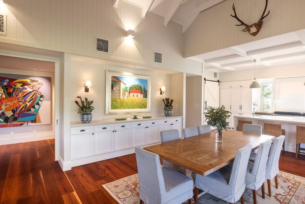 Bright, refined open-plan dining and kitchen interior with white vertical wall panelling, exposed painted beams, high sloped ceilings, dark polished timber flooring, and a large timber dining table surrounded by upholstered chairs. A white built-in sideboard, artwork, sconces, pendant lighting, and decorative objects create a coastal-traditional feel, while the adjacent kitchen features white cabinetry and light benchtops. An art-filled adjoining room is visible through a wide opening.