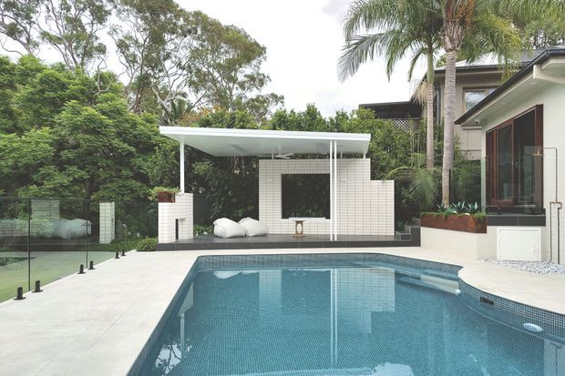 Contemporary outdoor pool area with a large tiled swimming pool, pale paved surrounds, and a minimalist white poolside pavilion or lounge structure. The pavilion features a flat roof, slim white columns, white tiled walls, a dark recessed opening, and built-in seating. Dense mature greenery and palm trees frame the property, with part of the modern house visible at right.