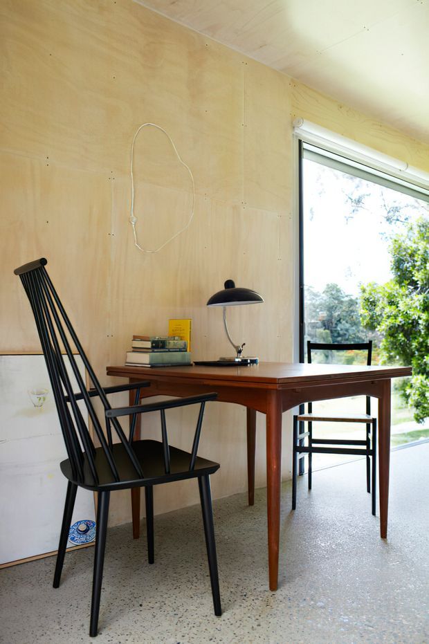 A bright, compact workspace or study nook with a mid-century timber desk, black spindle-back chair, desk lamp, books, and a second chair by a large window. The room has pale plywood wall and ceiling linings, a speckled terrazzo-like floor, and abundant natural light with greenery visible outside.