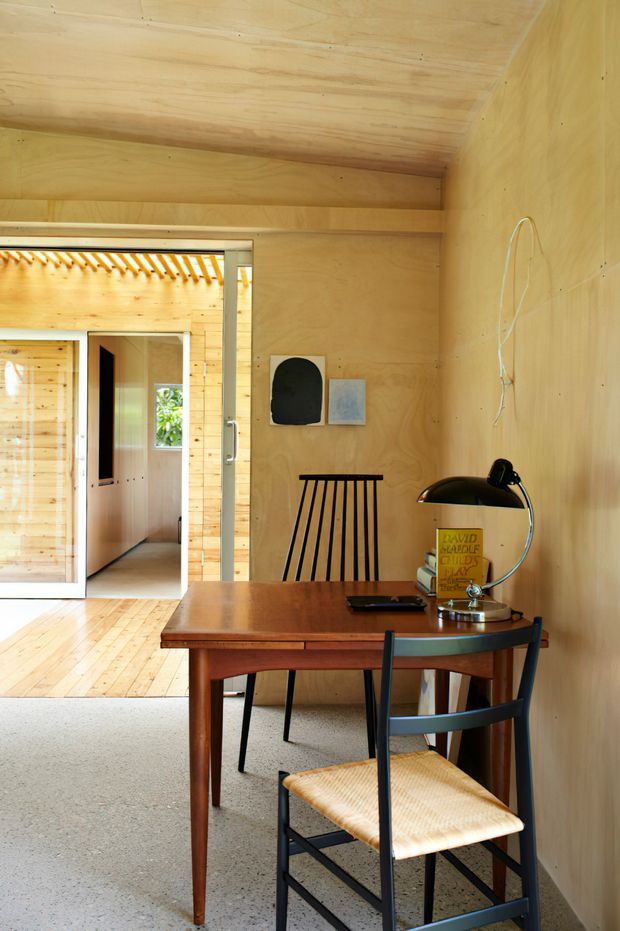 A compact wood-lined home office or study with warm plywood walls and ceiling, a timber writing desk, black-framed woven-seat chair, matching spindle-back chair, and a vintage-style desk lamp. A glazed sliding doorway opens to a timber-lined corridor and adjacent room, with daylight filtering through a slatted pergola or screen beyond.
