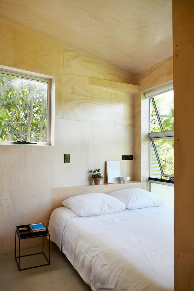 A compact, light-filled bedroom with plywood-lined walls and ceiling, a simple built-in timber bedhead shelf, and a white bed. Large opening windows bring in greenery, while a small black metal bedside table and minimal accessories create a pared-back, natural interior.