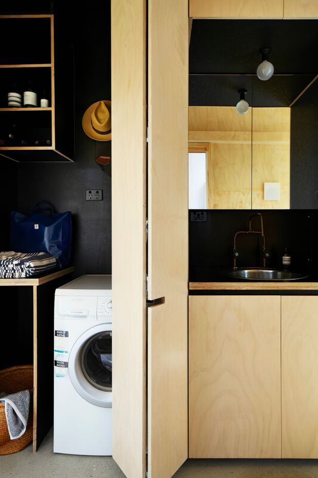Compact integrated laundry and kitchenette viewed through an open timber door. Pale plywood cabinetry and wall panels contrast with black walls, shelving, splashback, and ceiling panels. A front-loading washing machine, sink with dark tapware, mirrored upper cabinetry, open shelving, bench seating, and storage are visible. The space has a warm, functional, minimalist fit-out with strong black-and-light-wood contrast.