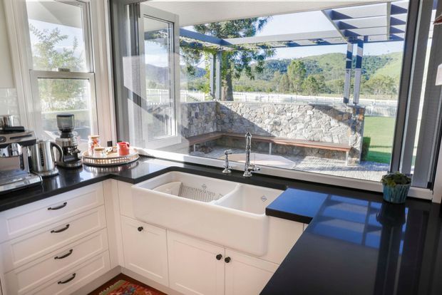 Bright white kitchen centred on a deep farmhouse-style apron-front sink beneath large windows and an open glazed door. White shaker cabinetry with dark hardware is paired with glossy black benchtops, while a coffee station occupies the left counter. Through the glazing is a covered outdoor entertaining area with a pergola, stone retaining or fireplace wall, built-in bench seating, lawn and wooded hills beyond.