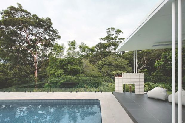 A contemporary pool terrace set within dense greenery. The image shows a rectangular swimming pool, pale paved decking, dark tiled outdoor flooring, a slim white flat-roofed pavilion with slender columns, a small white masonry feature wall, and large white outdoor cushions. A glass balustrade separates the terrace from the leafy landscape.