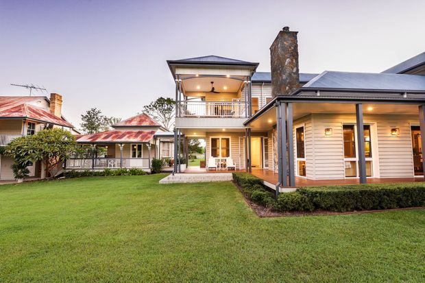 Exterior view of a substantial multi-wing residential property surrounding a broad manicured lawn. The building features light horizontal weatherboard cladding, dark metal hipped roof sections, covered verandas and balconies, timber-framed doors and windows, and a prominent stone-clad chimney. Older detached-looking wings with weathered red metal roofs connect to newer or renovated sections, creating a traditional Australian homestead character.
