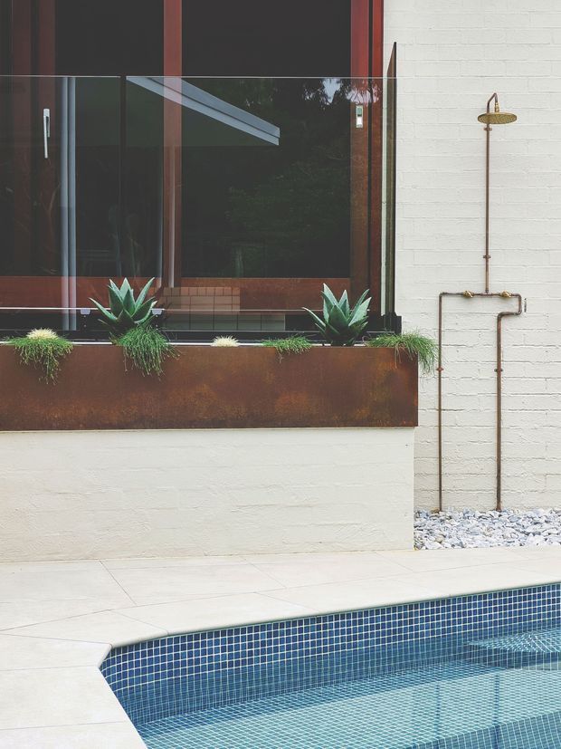 A contemporary outdoor courtyard with a tiled swimming pool in the foreground, pale paving, a white-painted brick wall, and an exposed copper outdoor shower. A rusted corten-steel planter beneath dark-framed glazing contains sculptural succulents and grasses, creating a refined industrial-modern landscape.