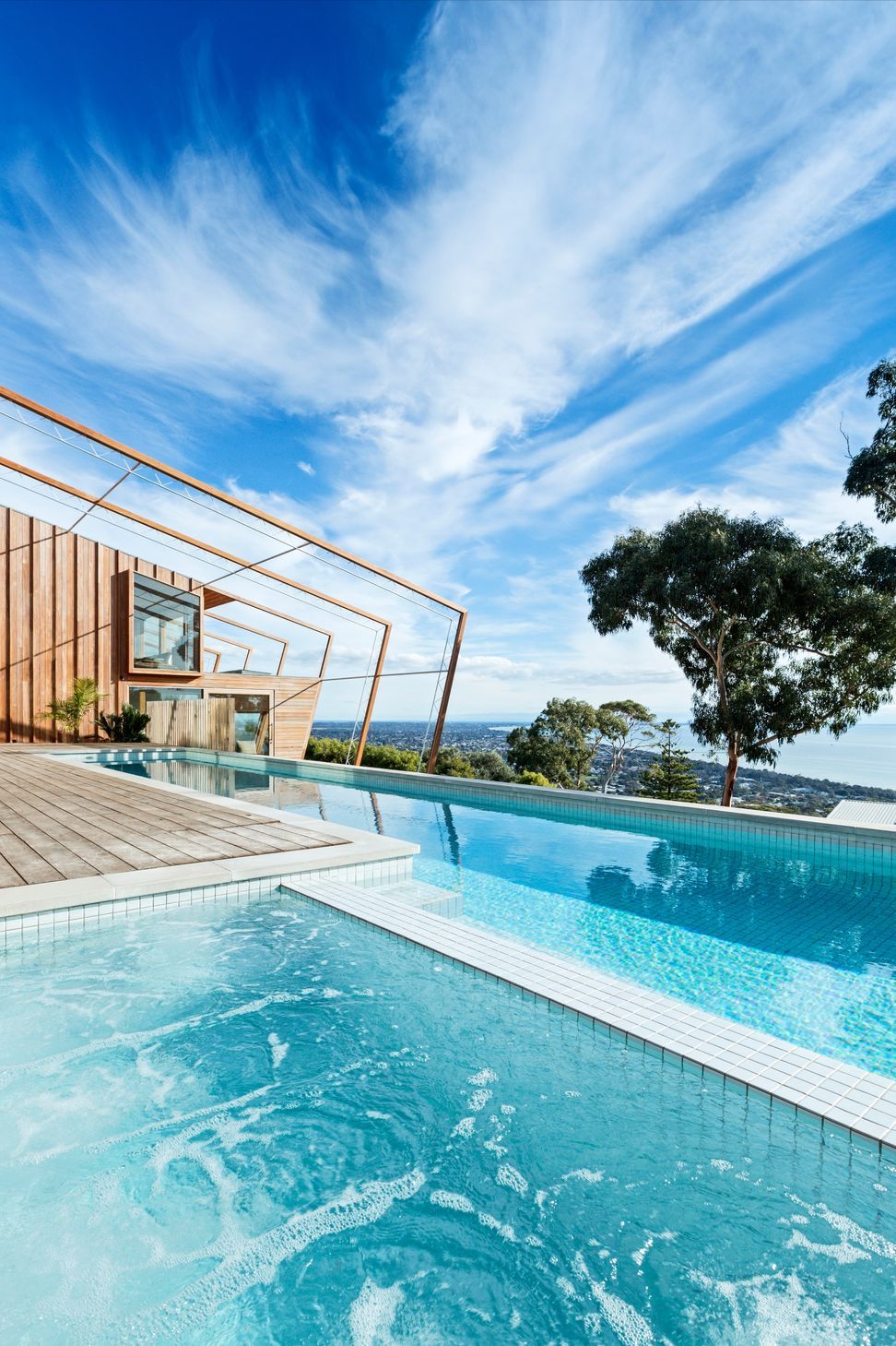 A white-tiled lap pool and integrated spa sit adjacent to an expansive timber deck, framed by striking angled timber trellis portals