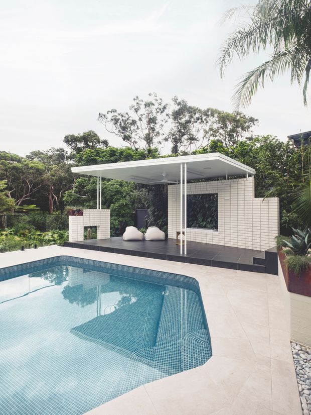 A modern outdoor pool area set within lush tropical landscaping. The foreground features a large curved swimming pool with blue mosaic tiling and pale stone paving. Behind it is a minimalist covered outdoor lounge or pavilion with a thin flat roof, slender white columns, white grid-pattern wall tiles, dark flooring, built-in bench seating, and pale cushions. Palm foliage and dense greenery surround the space.