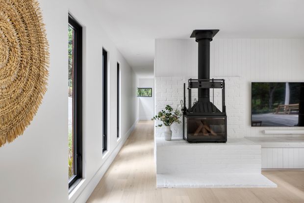 Bright contemporary open-plan living area with a white-painted brick fireplace surround and black freestanding wood-burning stove, pale timber flooring, black-framed windows, vertical white wall panelling, and a wall-mounted television. A hallway and glazing extend along the left side, while a woven circular wall feature adds natural texture.