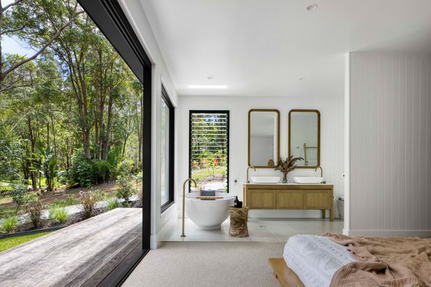 Bright, refined bedroom with an open-plan ensuite bathroom. The room features a freestanding white bathtub beside full-height black-framed glazing, a timber vanity with twin vessel basins, rounded brass-framed mirrors, white vertical wall panelling, pale carpet, and a timber bed partially visible in the foreground. The glazing opens toward a leafy garden and deck.