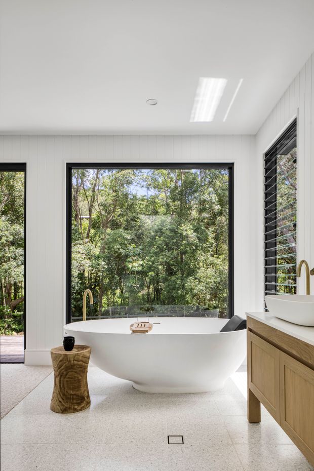 Bright, contemporary bathroom featuring a freestanding white bathtub positioned in front of expansive black-framed glazing overlooking dense greenery. The room has white vertical wall panelling, pale terrazzo-style flooring, a timber vanity with a white vessel basin, dark louvred window panels, and a sculptural timber stool.