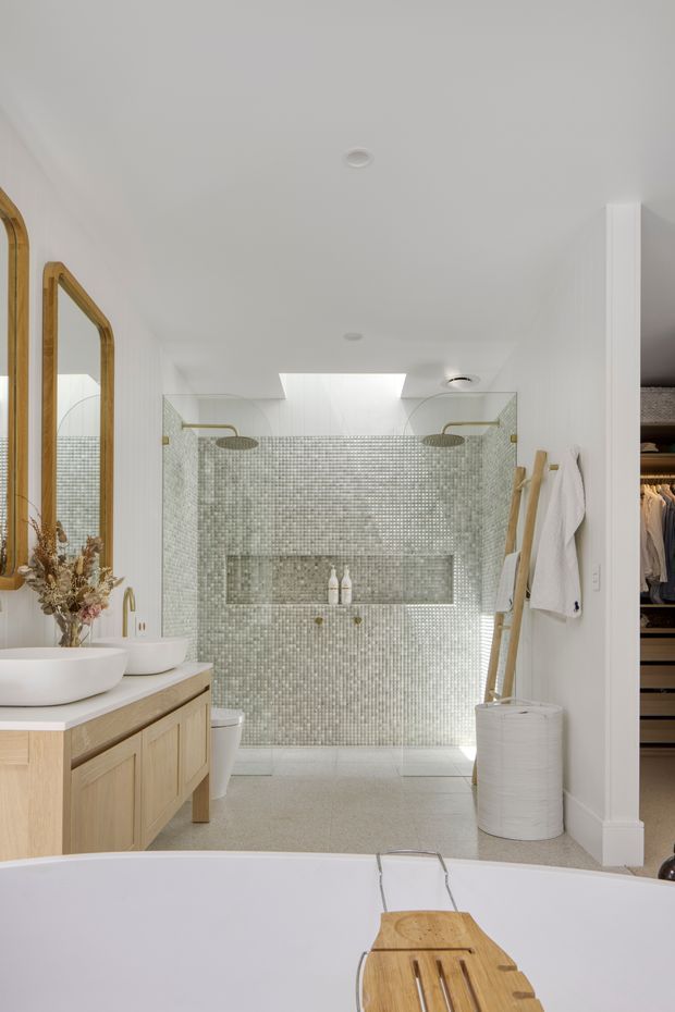 Bright, refined bathroom with a freestanding bathtub in the foreground, a pale timber double vanity with white vessel basins, a large timber-framed mirror, and a walk-in dual shower clad in small pale green-grey mosaic tiles. Brass shower fittings, white walls, a skylight, recessed lighting, open shelving, towels, and a partially visible walk-in wardrobe contribute to the calm, natural design.