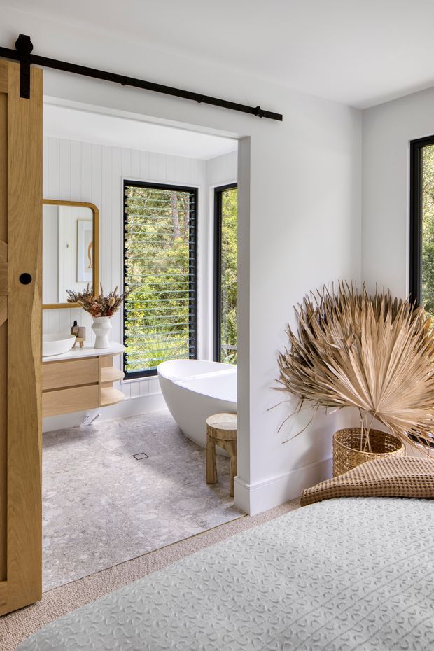 Bright contemporary bathroom viewed through an open timber barn-style sliding door from an adjacent bedroom. The bathroom features a freestanding white bathtub, wall-mounted timber vanity with a white basin, black-framed louvred windows, white vertical wall panelling, and a pale stone-look floor. Natural dried foliage and woven accessories add a warm organic accent.