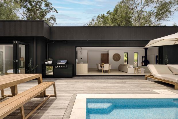 A contemporary single-level home opening onto a pool terrace. The dark vertically ribbed exterior and broad roofline frame an open-plan interior with a pale kitchen, dining area and living room. A timber outdoor dining table, built-in barbecue, sun loungers and large umbrella occupy the deck surrounding the swimming pool. Mature trees are visible beyond the property.