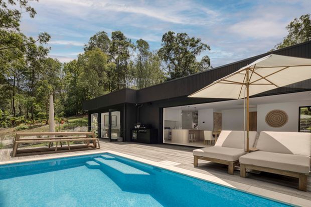 A contemporary single-storey house with a dark, angular roofline and extensive glazing, opening onto a timber deck and bright blue swimming pool. The outdoor entertaining area includes a timber picnic table, upholstered sun loungers, and a large cream umbrella, with established trees surrounding the property.