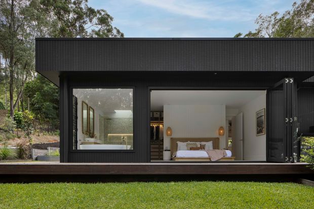 A compact, dark-clad contemporary pavilion set within a leafy landscaped setting. The open facade reveals a bedroom with a central bed, pendant bedside lights, built-in storage, and an adjacent bathroom visible through glazing. A broad timber deck runs along the front.