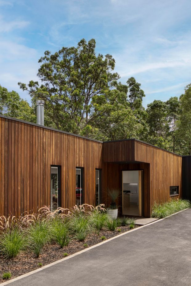 Contemporary single-storey building exterior clad in vertical warm timber boards, with a low-pitched roofline, dark-framed windows and recessed entry. Native trees and ornamental grasses surround the structure, while a smooth paved driveway runs along the foreground.