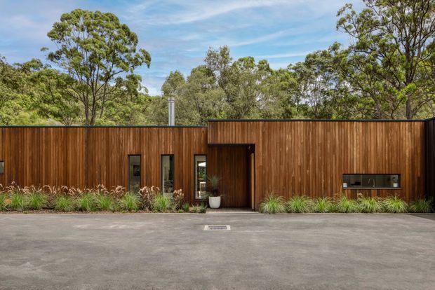 Single-storey modern building facade clad in vertical warm timber, set within a leafy natural landscape. The long, low frontage has narrow windows, a recessed entry, a dark-framed built-in opening at the right, landscaped planting along the base, and a broad paved foreground.