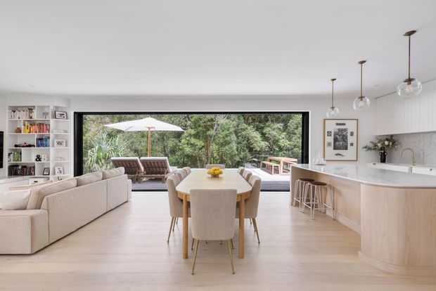 Open-plan contemporary kitchen, dining and living space with pale timber flooring, a large light-wood kitchen island, white cabinetry, integrated shelving, and a neutral upholstered dining and lounge setting. Full-width black-framed glazing opens the room visually to a leafy outdoor area, while globe pendant lights hang above the island and dining zone.