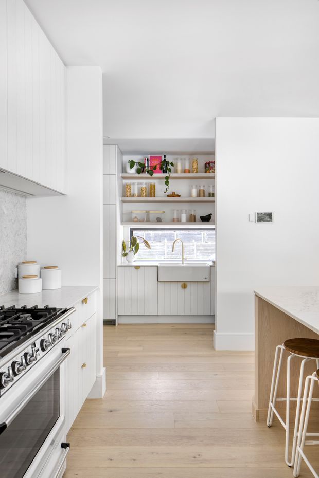 Bright, predominantly white kitchen with shaker-style cabinetry, a large freestanding range, open display shelving, a farmhouse-style sink beneath a window, and a light timber island with stools. Pale timber flooring and minimal decorative accents create a clean, warm contemporary look.