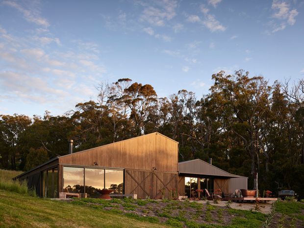 A low-set timber-clad rural dwelling or barn-style residence viewed from the landscaped grounds. The building has a prominent gable-ended central volume, dark pitched-roof wings, expansive glazed openings, and large timber barn doors. It is surrounded by mature woodland and sloping grassed terrain, with warm evening light reflecting in the glazing.