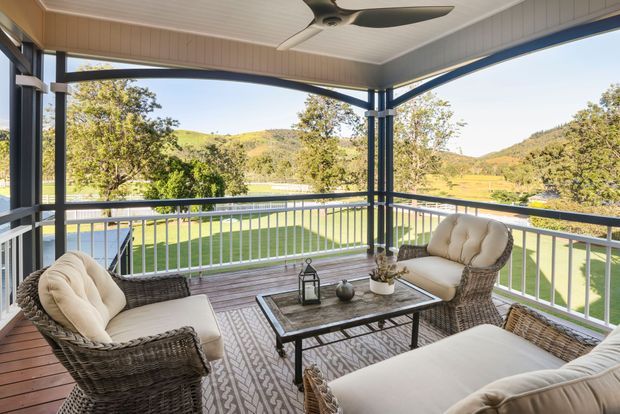 Covered and screened outdoor terrace with wicker lounge seating, a coffee table, ceiling fan, and white balustrade overlooking a landscaped lawn and rolling hills. The space has a relaxed, airy resort-like character with dark structural framing and timber-look decking.
