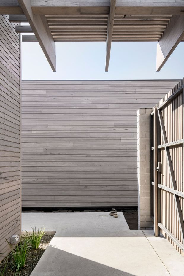 A minimalist architectural courtyard or entry passage enclosed by pale horizontal timber-clad walls and a slatted timber pergola. The space features a smooth concrete walkway and courtyard floor, a tall slatted gate on the right, a small planted garden bed, and subtle stacked-stone landscaping. Strong sunlight creates geometric shadows across the paving and emphasizes the exposed timber structure.