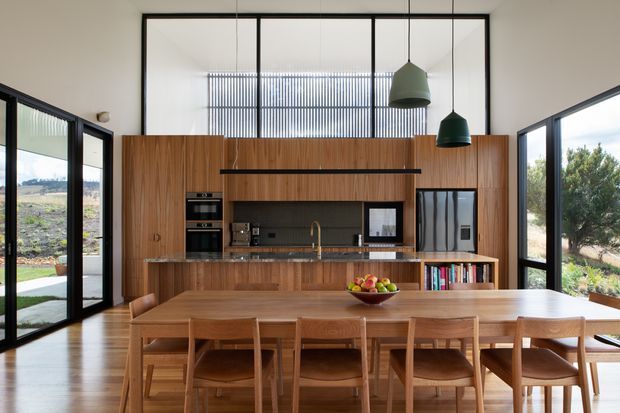 A spacious open-plan kitchen and dining area with full-height glazing, warm timber cabinetry and flooring, and a long timber dining table surrounded by chairs. The kitchen features integrated appliances, a dark benchtop and splashback, open shelving, and three pendant lights. Black-framed glass doors and windows provide views to the surrounding landscape.
