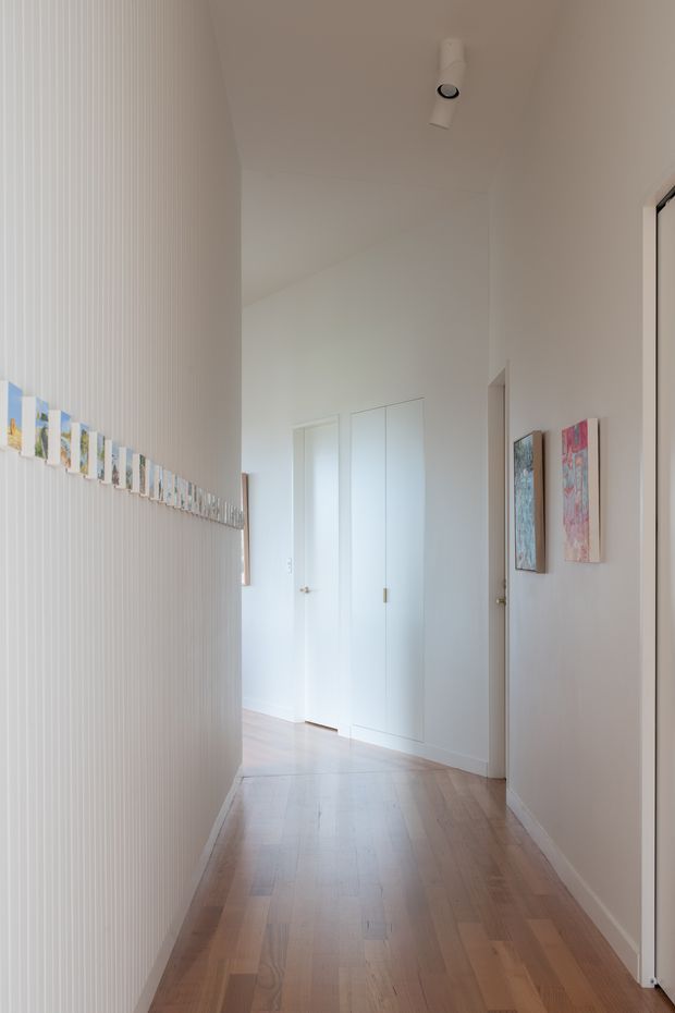 A bright, narrow residential hallway with white walls and vertical ribbed wall panelling on the left, displaying a horizontal line of small artworks or photographs. The hall has pale timber flooring, white flush doors and built-in cupboards, simple white ceiling track lighting, and additional colourful artwork on the right wall. The space is clean, minimal, and softly lit.