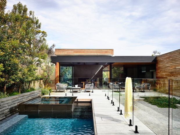 Exterior view of a low, modern house opening onto a landscaped courtyard and swimming pool. The building features dark horizontal roof planes, warm timber-clad walls, extensive floor-to-ceiling glazing, and a timber pool deck with frameless glass safety balustrades. Outdoor lounge furniture sits beneath the covered terrace, while concrete and gravel landscaping frame the pool.