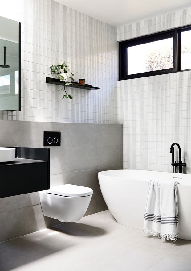 A bright, contemporary bathroom featuring a freestanding white bathtub, wall-hung toilet, black floating vanity with white basin, and matte black tapware. The walls are clad in fine vertically ribbed white tiles, with larger grey concrete-look tiles around the toilet and bath. A black-framed horizontal window, black-framed mirror, floating shelf, and trailing plant add contrast. The room has a clean, refined, monochromatic appearance.
