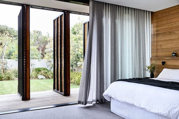 A bright contemporary bedroom opening onto a landscaped garden through large glazed folding doors with dark timber frames and external louvred screens. The room features a low-profile bed with white bedding, a grey carpet, full-height grey curtains, and a warm horizontal timber feature wall with minimal bedside shelving.