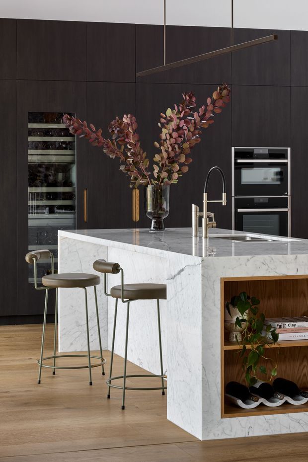 A refined contemporary kitchen centred on a large white-veined marble island with an integrated sink and tall curved tap. The island includes an open dark timber display niche with books, plants and wine storage. Behind it, full-height dark timber cabinetry conceals integrated appliances, including a wine refrigerator and stacked ovens. Two slim upholstered bar stools sit at the island, while a linear suspended pendant light spans the space. Warm timber flooring and deep brown cabinetry create a sophisticated, moody atmosphere.