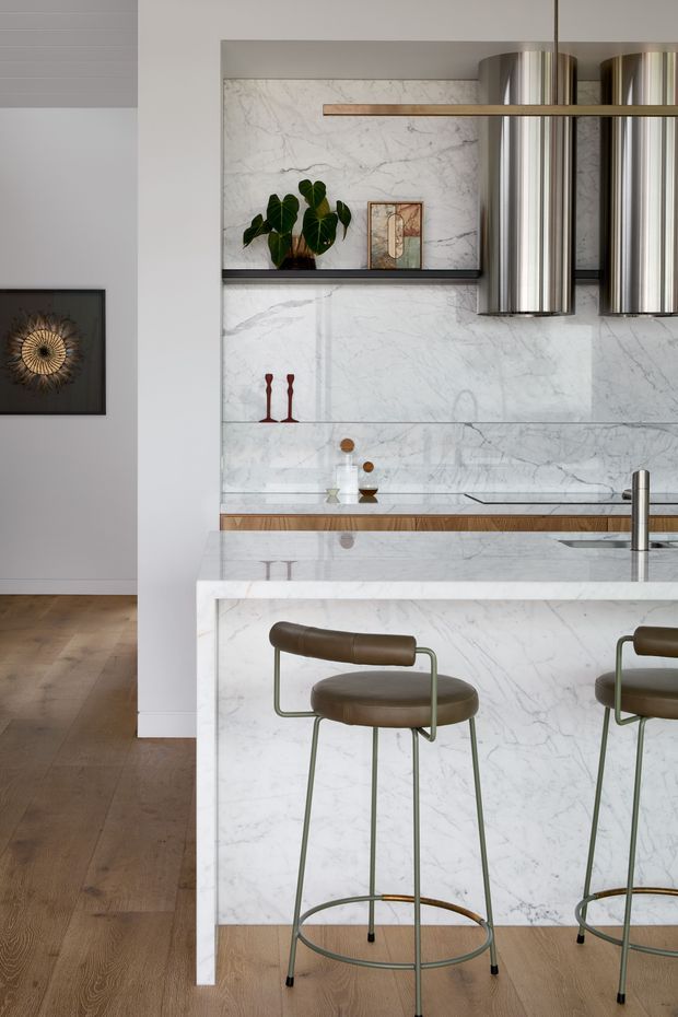 Refined contemporary kitchen with extensive white veined marble or stone surfaces, including a full-height splashback, island waterfall edge and benchtop. The kitchen features a timber island detail, brushed metal cylindrical rangehoods, an undermount sink with a slim metal tap, and two sculptural brown leather bar stools with slender green-grey metal frames. Warm timber flooring and neutral painted walls create a balanced, sophisticated interior.