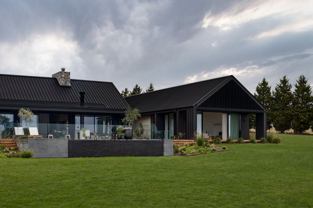 A contemporary single-storey home set in a landscaped lawn, composed of dark vertically clad gabled volumes with steep metal roofs. Large glazed openings connect the living areas to a terrace and raised garden beds, while a stone chimney and mature evergreen planting add contrast. The image is an exterior architectural photograph under an overcast sky.
