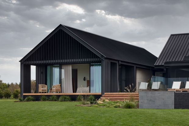 Exterior view of a contemporary single-storey house with a dark vertically clad gable-roof pavilion, broad glazed openings, timber decking, and a connected wing with matching dark roofing. The home sits beside a manicured lawn and landscaped garden under an overcast sky.