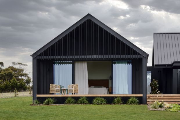 Exterior view of a compact modern gabled pavilion or cabin with dark vertical cladding and a strongly expressed triangular gable facade. The front is largely open through floor-to-ceiling glazing or doors, revealing a bedroom with a bed, pale curtains, and two woven chairs. A timber deck runs along the front, bordered by low ornamental planting and lawn. A second dark-clad gabled structure is visible to the right under an overcast sky.