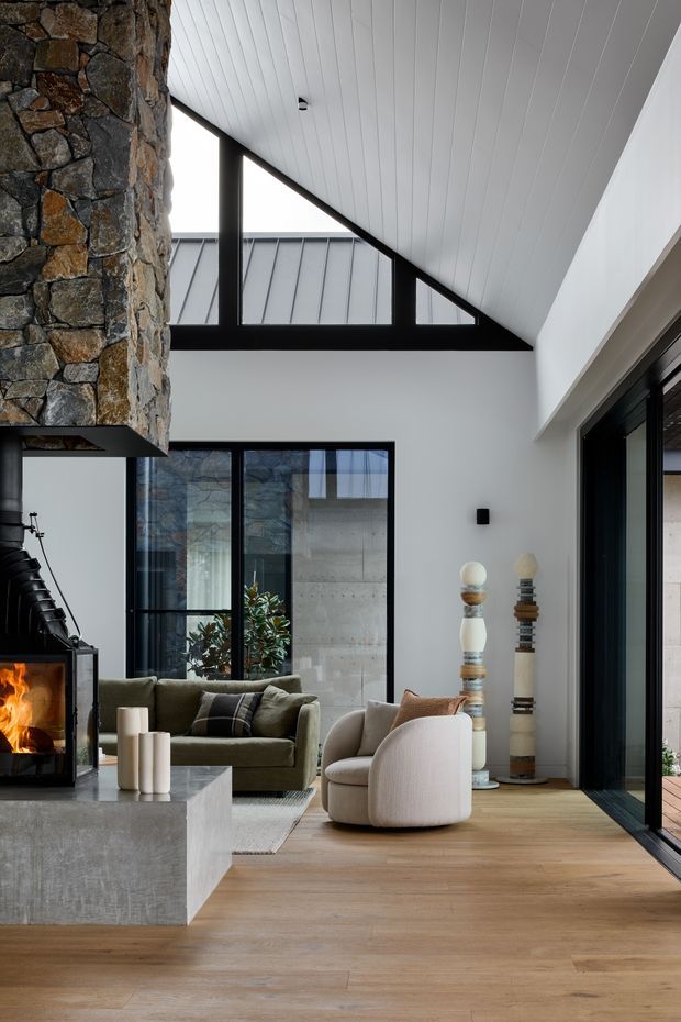 A refined contemporary living room with a high sloping ceiling and clerestory glazing. The room features a dramatic rough-stone fireplace wall, black-framed windows and doors, white vertical-panel ceiling, pale walls, warm timber flooring, a concrete hearth or coffee table, and soft neutral seating. A lit fire and sculptural decorative objects create a warm, sophisticated atmosphere.