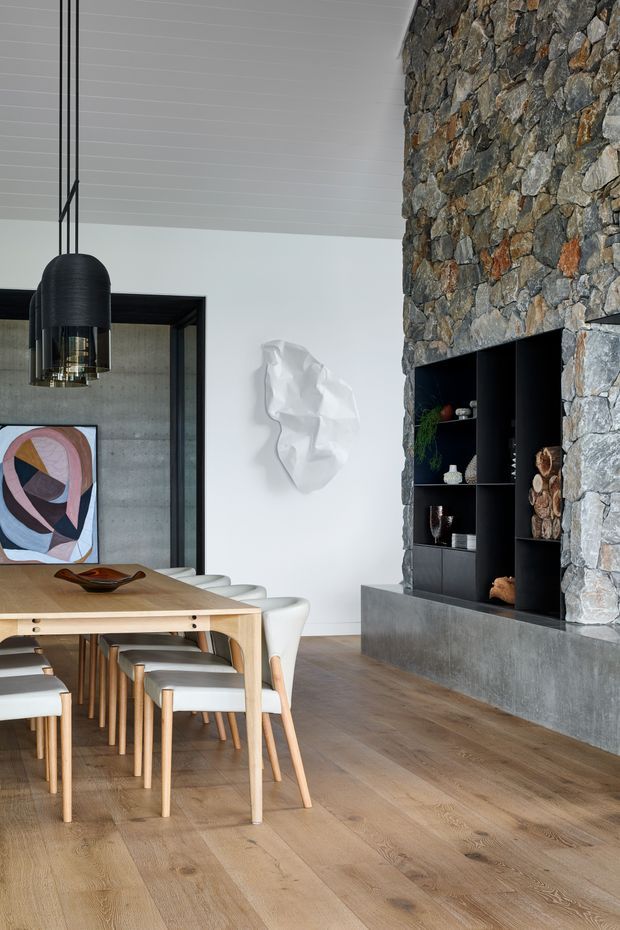 Contemporary dining area with a large light timber dining table and upholstered chairs, set against a white wall and a striking rough natural-stone feature wall. A black built-in shelving unit and concrete hearth or plinth occupy the stone wall, while a large black pendant light hangs above the table. Timber flooring, framed glazing, artwork, and sculptural wall decor complete the refined interior.