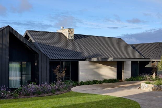 A contemporary single-storey residential building with dark vertical cladding and intersecting pitched metal roofs. Large glazed openings, a stone chimney, sheltered entry, landscaped garden beds, and a curved paved driveway are visible under a blue evening sky.