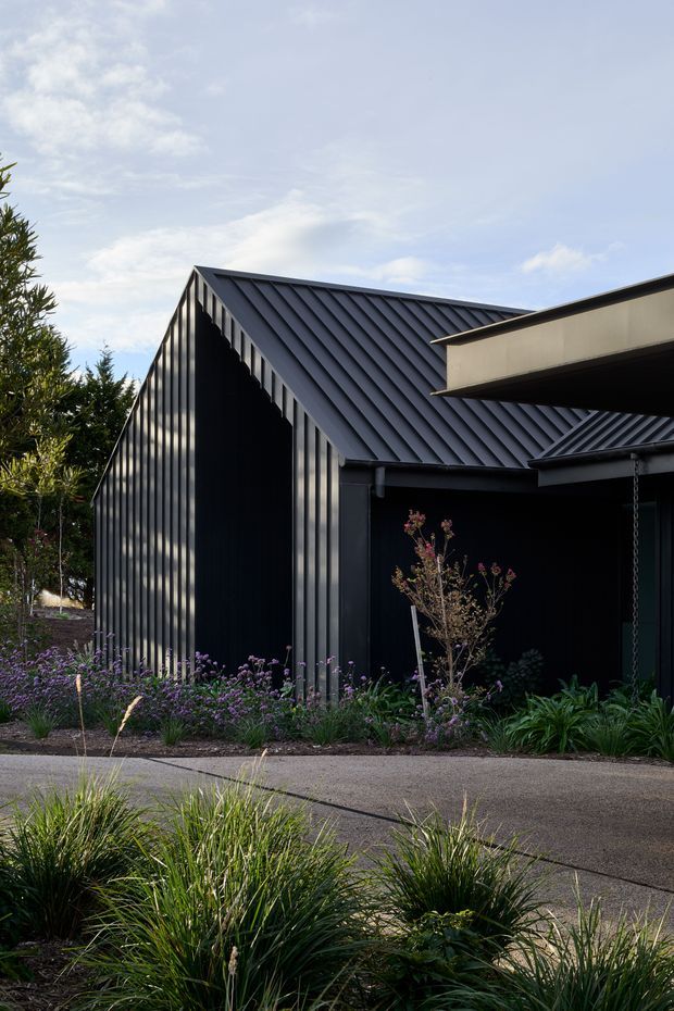 Exterior view of a contemporary dark-clad building with a prominent steep gable roof and standing-seam metal roofing. The facade combines vertical dark metal cladding with deep shadowed openings and a projecting flat-roofed element. Low ornamental planting and a paved pedestrian path occupy the foreground.