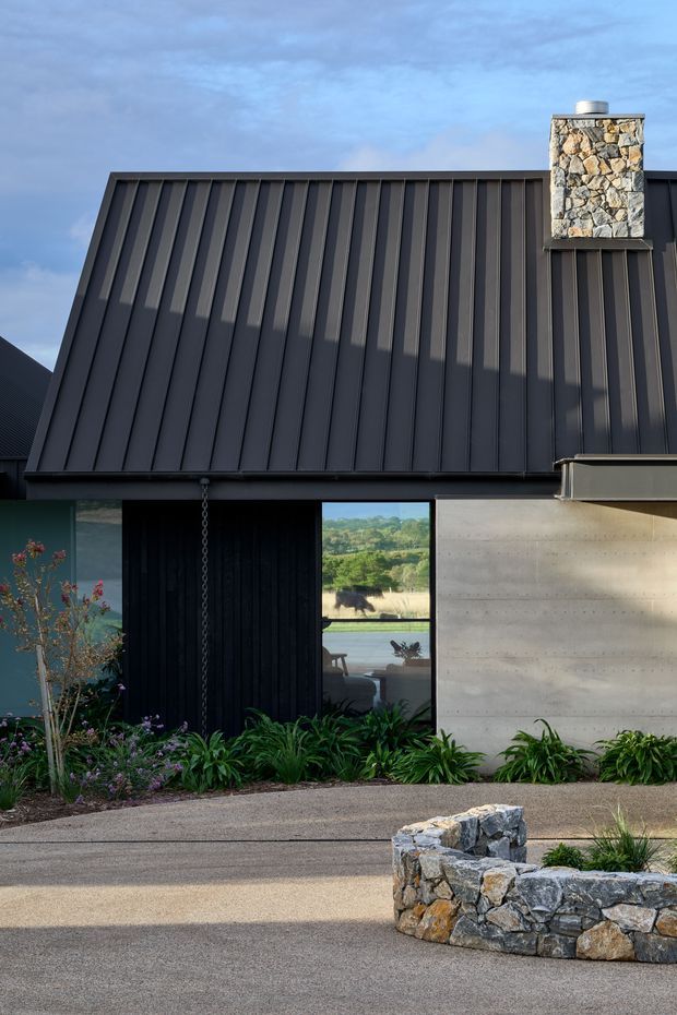 Exterior view of a contemporary single-storey building with a steeply sloping dark standing-seam metal roof and matching dark vertical cladding. A prominent rough stone chimney rises above the roof, while a smooth pale concrete wall and large glazed opening form the lower facade. Native-style planting and low stone retaining walls frame the paved foreground.
