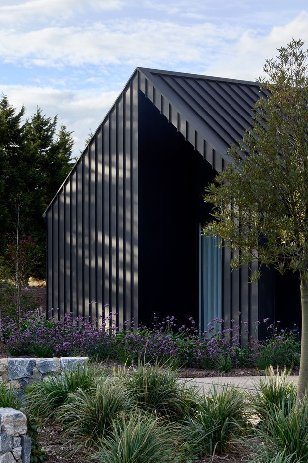Exterior view of a contemporary dark-clad building with a steep gabled roof and strong vertical ribbed facade. The structure is set within a landscaped garden featuring ornamental grasses, purple flowering plants, trees, stone edging and a paved walkway. A tall blue-toned glazed doorway is partially visible beneath the deep gable.