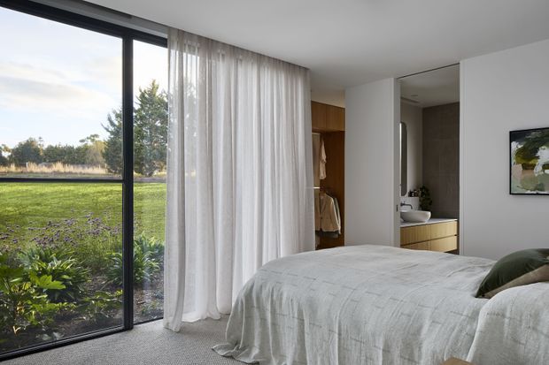 Bright contemporary bedroom with a large black-framed floor-to-ceiling window overlooking a lawn and garden. Sheer white curtains, a textured light bedcover, built-in timber wardrobe storage, and an adjoining open vanity and bathroom area create a calm, refined interior.