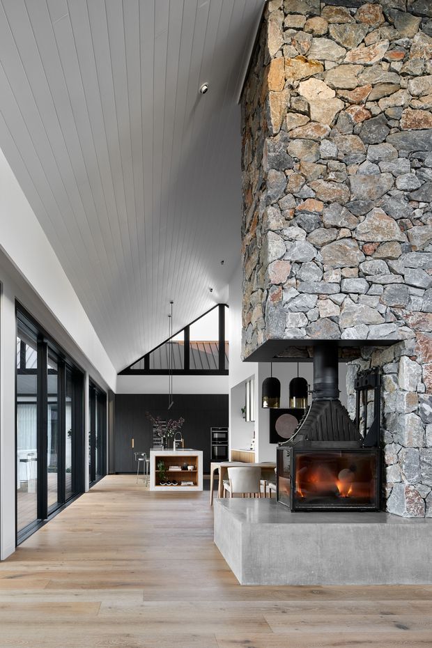 A contemporary open-plan living and kitchen interior with a dramatic floor-to-ceiling rough stone fireplace and black wood-burning stove set on a concrete hearth. The room features pale timber flooring, a white linear-panel ceiling, extensive black-framed glazing along one side, dark cabinetry, a dining area, and a high glazed gable-like opening that creates an airy, elongated volume.