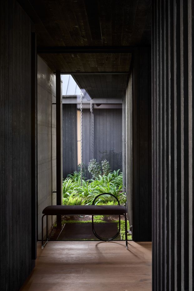 A dark, narrow covered passage or entryway opens onto a lush planted courtyard. The space features vertically grooved dark wall cladding, a timber-plank ceiling, warm timber flooring, exposed structural framing, and a simple dark bench with a curved metal support. Soft daylight filters in from the courtyard, creating a dramatic contrast between the moody interior and greenery beyond.