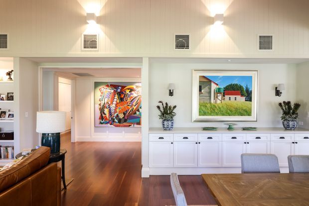 Bright, refined open-plan dining and living area with white built-in cabinetry, a large timber dining table, leather seating, polished timber flooring, and high vertical wall panelling. Artwork, decorative objects, and wall sconces add colour and character, while a wide opening connects to an adjoining living or gallery space.