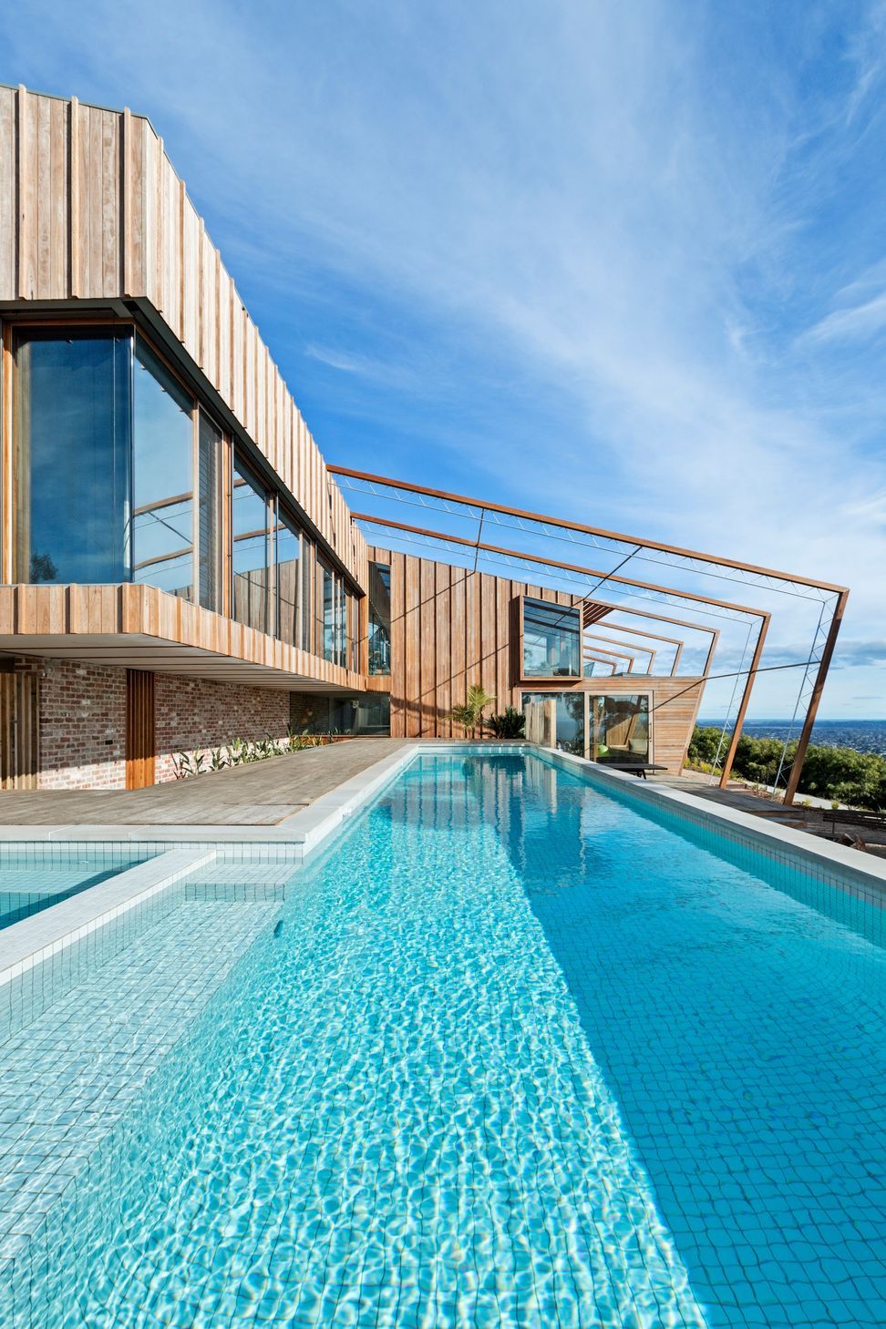 A white-tiled lap pool integrates with the timber deck, beneath vertical hardwood cladding and a structural pergola framing the horizon