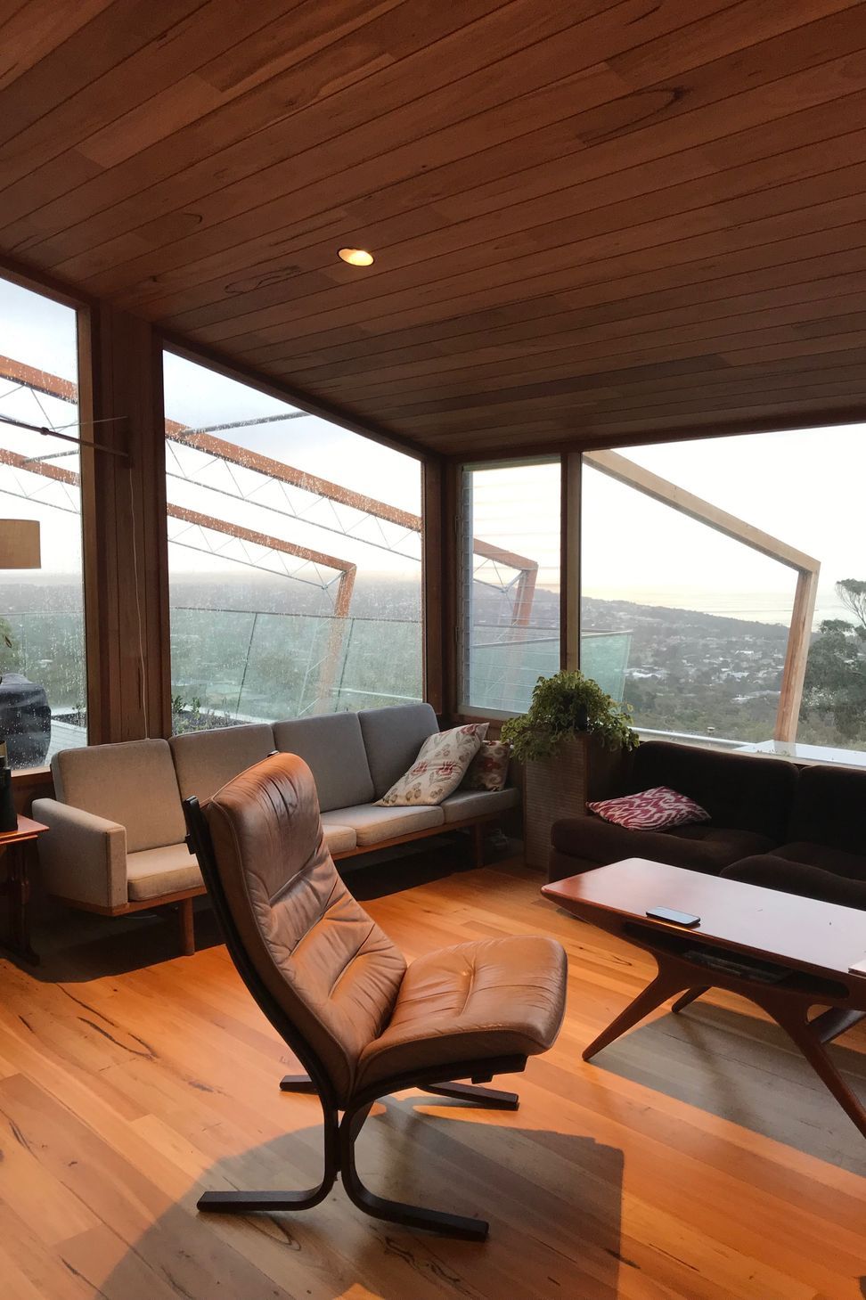 Continuous timber-lined ceilings and warm hardwood floors create a refined, mid-century inspired living space that looks out to panoramic coastal views