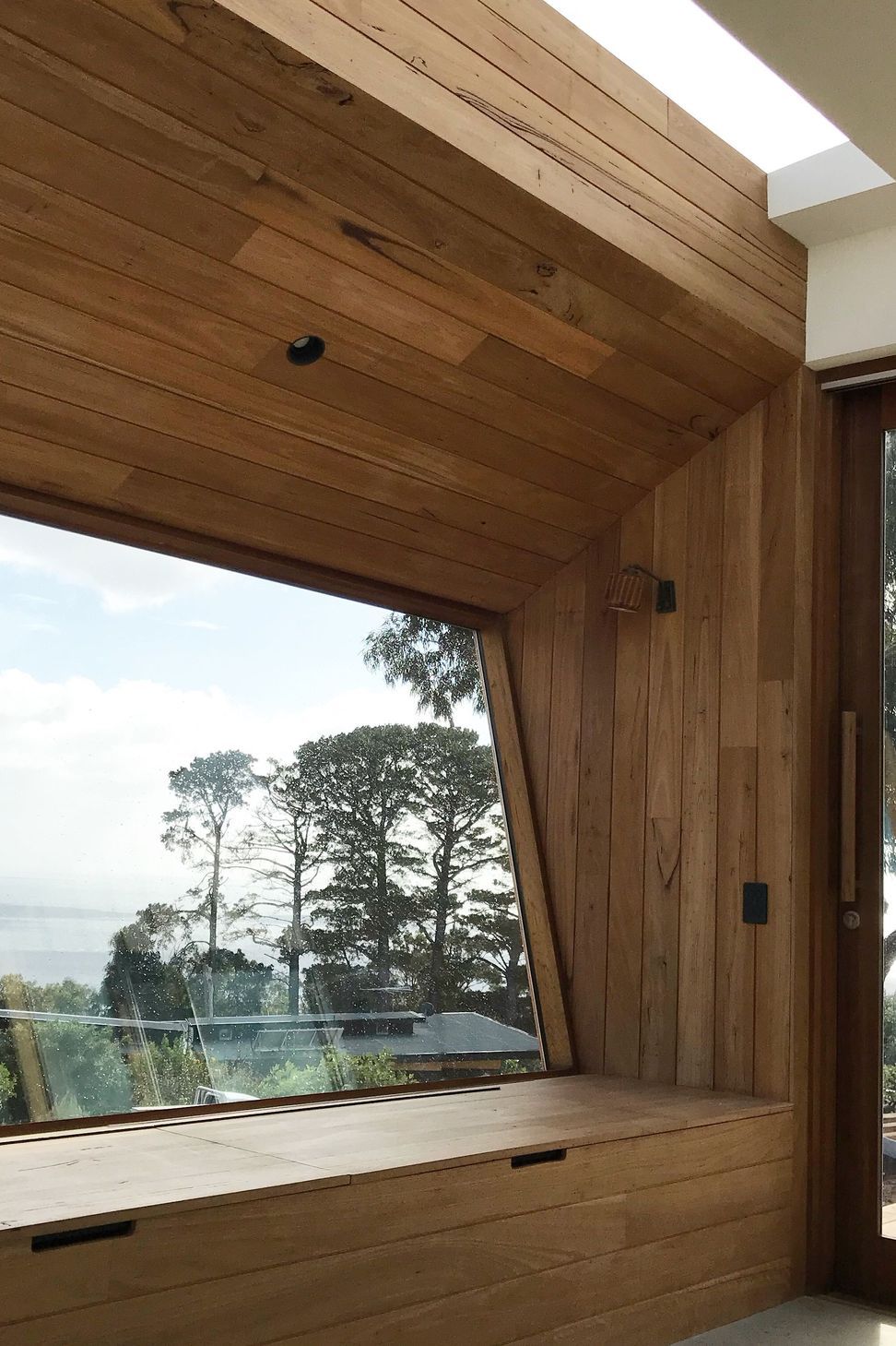 A custom timber window daybed sits beneath a raked ceiling and an integrated skylight, framing an angled view down the slope of the coastal landscape